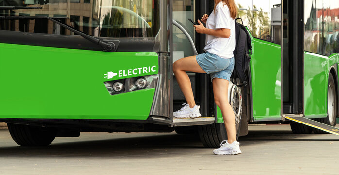 Female passenger gets on the green coloured electric bus at a bus stop in city in summer.
