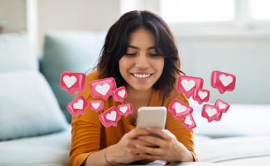 Middle eastern woman lies on a bed, looking down at a smartphone in her hands. She is smiling and appears happy. Above her phone, there are digital heart icons representing likes