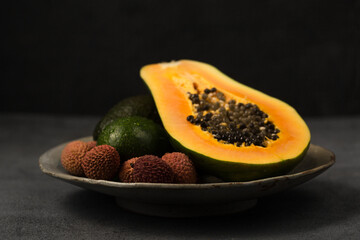Two avocados, half a papaya and lychee berries on plate made by hand on a gray background