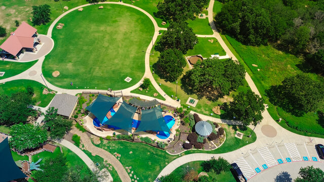 Suburban park with pavilion shelter, slash pad sail shade structure and diverse kids playing water in large modern children recreation center, lush green trees in Flower Mound, Texas, aerial view