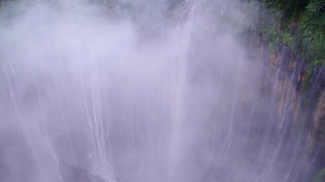 Aerial view of misty Tumpak Sewu waterfall surrounded by lush jungle and river, East Java, Indonesia.