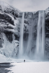 A lone hiker in front of massive majestic waterfall Skogafoss in south Iceland, on a winter morning, with snow and ice in the gorge, travel destination famous landmark iconic natural wonder