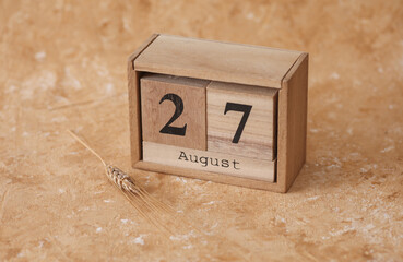 Wooden block calendar with the date August 27 and spikelets of wheat on a beige concrete background