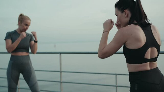 Focused female athletes engage in a boxing workout by a serene waterfront, demonstrating strength and teamwork.