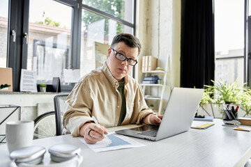 A young man with Down syndrome works on a laptop and paperwork in a bright office.