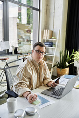 A young man with Down syndrome works on his laptop and a sticky note, looking focused and determined.