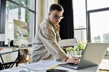 A young man with Down syndrome works on a laptop in a brightly lit office.