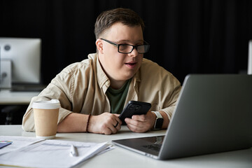 A young man with Down syndrome sits at a desk, working on his laptop while holding a smartphone.