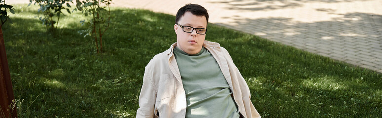 A young man with Down syndrome sits on a bench in a park, wearing glasses and a light jacket.