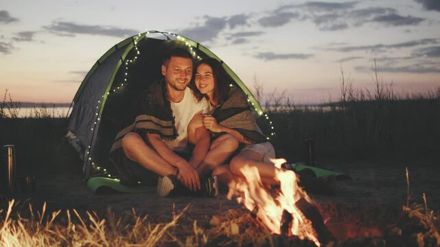 A couple is hugging next to their tent, both wrapped in a warm blanket and gazing at the campfire. Evening is turning into night, and string lights are hanging on the tent, creating a cozy atmosphere.