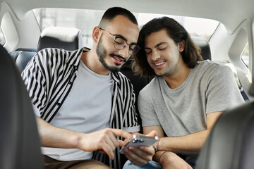 Two men share a smile and a phone screen in the backseat of a car.