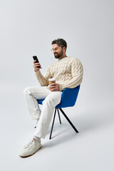 A bearded man in a white sweater sits peacefully in a chair, engrossed in his cell phone.