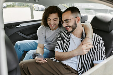 A gay couple sits in the backseat of a car, laughing and looking at a phone.