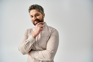 A stylish man with a beard poses in elegant attire against a grey background in a studio setting.