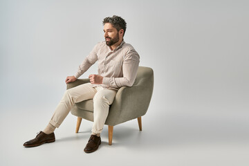 Bearded man in elegant attire sits with crossed legs in stylish pose on grey background in studio.