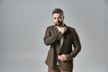 An alluring man with a beard strikes a pose in elegant attire against a grey studio background.