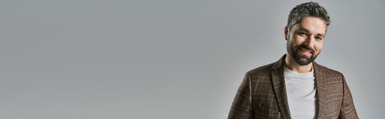 Obraz premium A stylish bearded man in elegant attire stands confidently in front of a sleek gray backdrop in a studio setting.