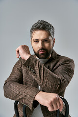 A bearded man in a brown jacket exudes style while holding a black bag against a grey background.