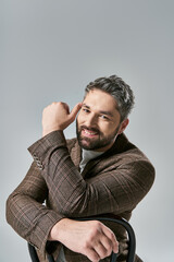 A stylish man with a beard sits in a chair, hand on head, lost in thought, exuding elegance and...