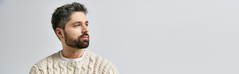 Obraz premium A charismatic man with a beard strikes a pose in a cozy white sweater against a grey studio background.