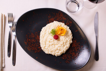 Risotto acquerello agli agrumi guarnito con una fetta d'arancia, rosmarino e una mora e servito in un piatto nero in un ristorante elegante