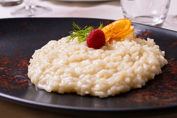 Risotto acquerello agli agrumi guarnito con una fetta d'arancia, rosmarino e una mora e servito in un piatto nero in un ristorante elegante