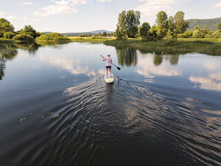 Woman paddling a white stand up paddle board exploring the lakeshore and enjoying a fantastic nature view, aerial shot. Health and wellness concepts.