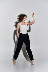 Fototapeta premium A pretty young woman strikes a graceful pose in black pants and white tank top against a grey studio backdrop.