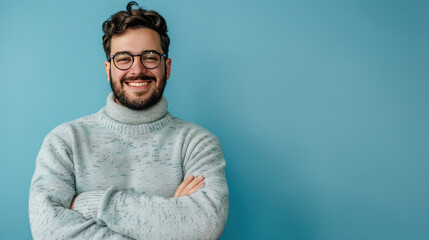 Obraz premium Young bearded man in a casual sweater and glasses, smiling happily with crossed arms, looking at the camera, blue backdrop