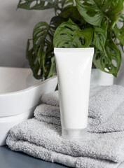 White cream tube on grey towels near vessel sink and green plant close up, cosmetic mockup