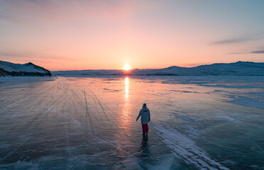 Fototapeta premium Aerial view on the young woman walking on the blue cracked ice of Baikal at beautiful orange sunrise. Sun reflections on the ice. Winter landscape of frozen Baikal