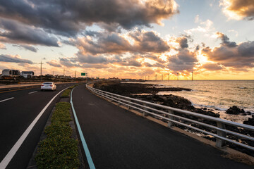 Hankyung, Jeju-si, Jeju-do, South Korea - October 11, 2019: Sunset view of a car driving on the road against sea rocks and wind generators on the sea at Sinchang Windmill Coast
