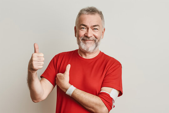 A senior man in a red shirt, smiling and showing thumbs up after donating blood with a bandaged arm, conveying a positive and supportive gesture