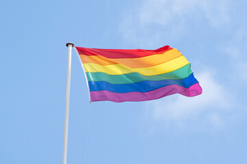 LGBTQ Rainbow Flag Flying From Pole Against Blue Sky To Celebrate Pride Month LGBTQIA