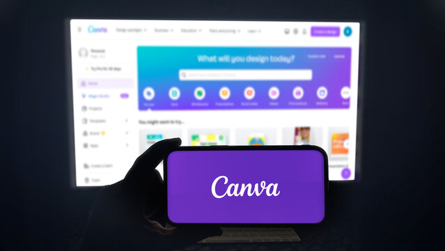Canva app running on a modern mobile phone. The smartphone is being held in landscape mode, in the darkness. A PC monitor with the Canva website in the background.