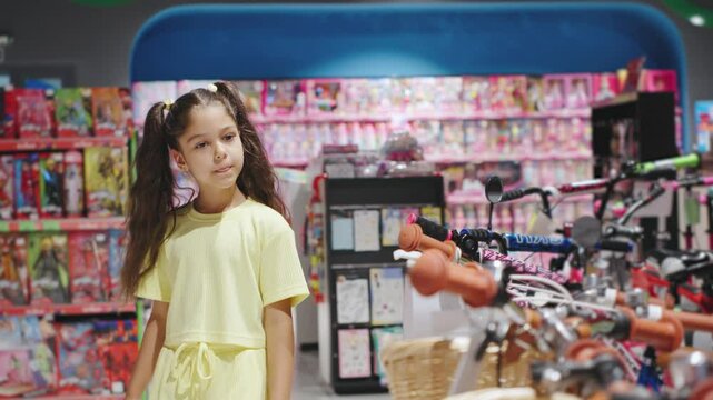 A little girl in a toy store is surrounded by dolls, bikes, and colorful items, creating an ambiance of joy and curiosity. Girl chooses a bicycle with a basket