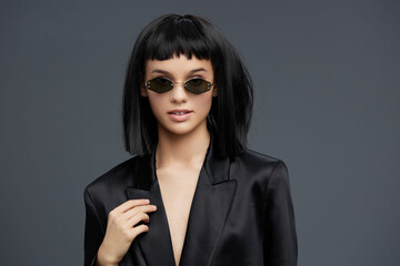 A stylish woman in a black suit and sunglasses confidently poses against a gray backdrop, embodying...