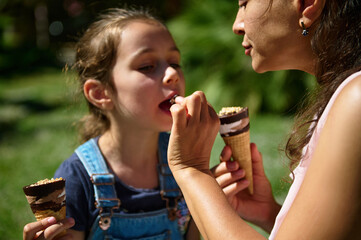 Mother feeding daughter ice cream in a sunny park, enjoying a sweet moment