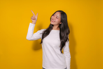 Young asian woman smiling and pointing in a studio with a yellow background, perfect for ads or marketing. Her inviting gesture suggests she's presenting a product or service