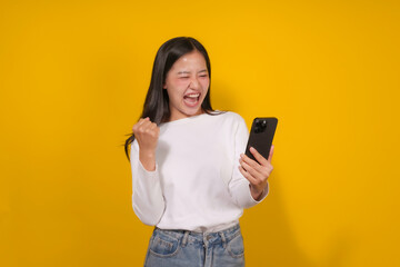 Fototapeta premium Young asian woman celebrates good news with a smartphone in hand, raising her fist in joy against a vibrant yellow background. Her beaming smile radiates success and happiness