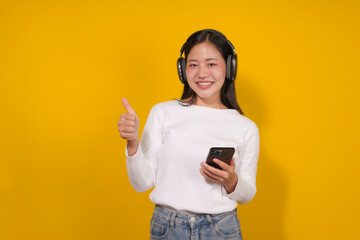 Young woman happily listens to music on her smartphone, giving a thumbs up against a vibrant yellow background