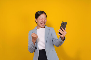 Young asian businesswoman holding a smartphone, raising her fist, and laughing, surprised by good news or business achievement