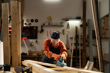 Female carpenter in safety glasses sanding wooden board using belt sander, working in her workshop.