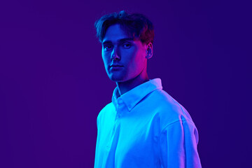 Portrait of young man, student wearing white shirt, standing with calm expression, looking at camera against gradient blue purple background in neon light. Concept of youth, human emotions, fashion