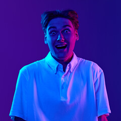Portrait of young man in white polo shirt standing with wide open eyes and mouth, expressing shock and excitement against gradient background in neon light. Concept of youth, human emotions, fashion
