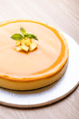 A beautifully glazed mango cheesecake topped with a dollop of mango puree and fresh mint leaves. The cake is presented on a silver cake board, capturing its smooth and shiny surface. 