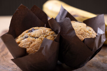 Freshly baked banana chocolate chip muffins wrapped in brown parchment paper, placed on a wooden cutting board alongside a bunch of ripe bananas. The rustic setup on a wooden table