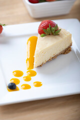 A slice of creamy cheesecake topped with a fresh strawberry, served on a white plate with a drizzle of fruit sauce. A bowl of fresh strawberries is in the background, adding vibrant color to the dish