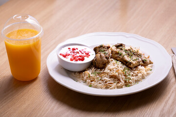 Delicious plate of biryani rice with tender chicken pieces, served with a side of yogurt garnished with pomegranate seeds, and accompanied by a refreshing orange juice in a plastic cup.