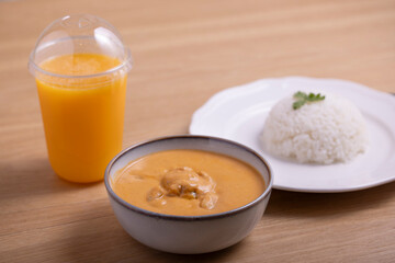 Delicious bowl of creamy butter chicken served with a mound of steamed white rice garnished with cilantro on a white plate, accompanied by a refreshing orange juice in a plastic cup. 